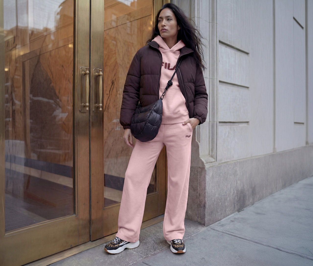 Femme portant une doudoune marron, un sweat à capuche et un pantalon de jogging roses, un sac matelassé noir et des baskets léopard devant un bâtiment.