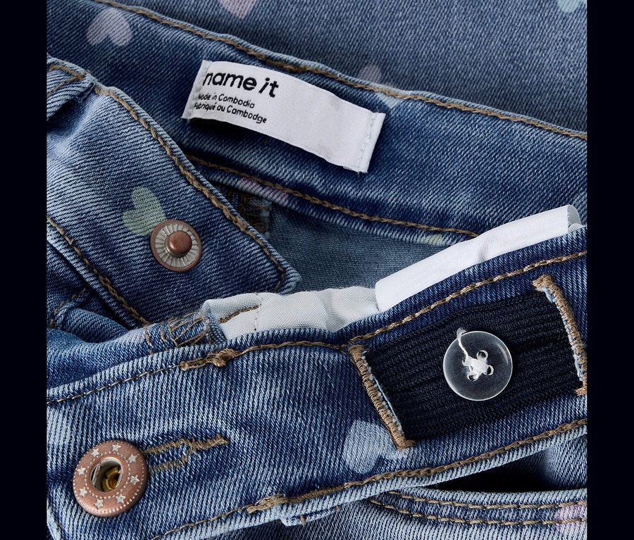 Gros plan sur un jean en denim bleu avec un motif de cœurs discret, montrant un bouton couleur cuivre, une étiquette "name it" et une taille élastique ajustable avec un bouton transparent.