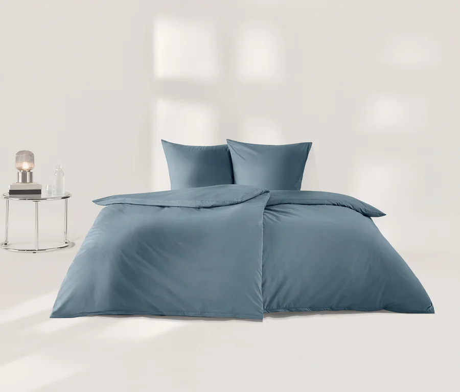 Parure de lit en percale bleue comprenant une housse de couette et deux taies d'oreiller, soigneusement disposée sur un sol clair, à côté d'une table de chevet chromée avec une lampe et une bouteille en verre.