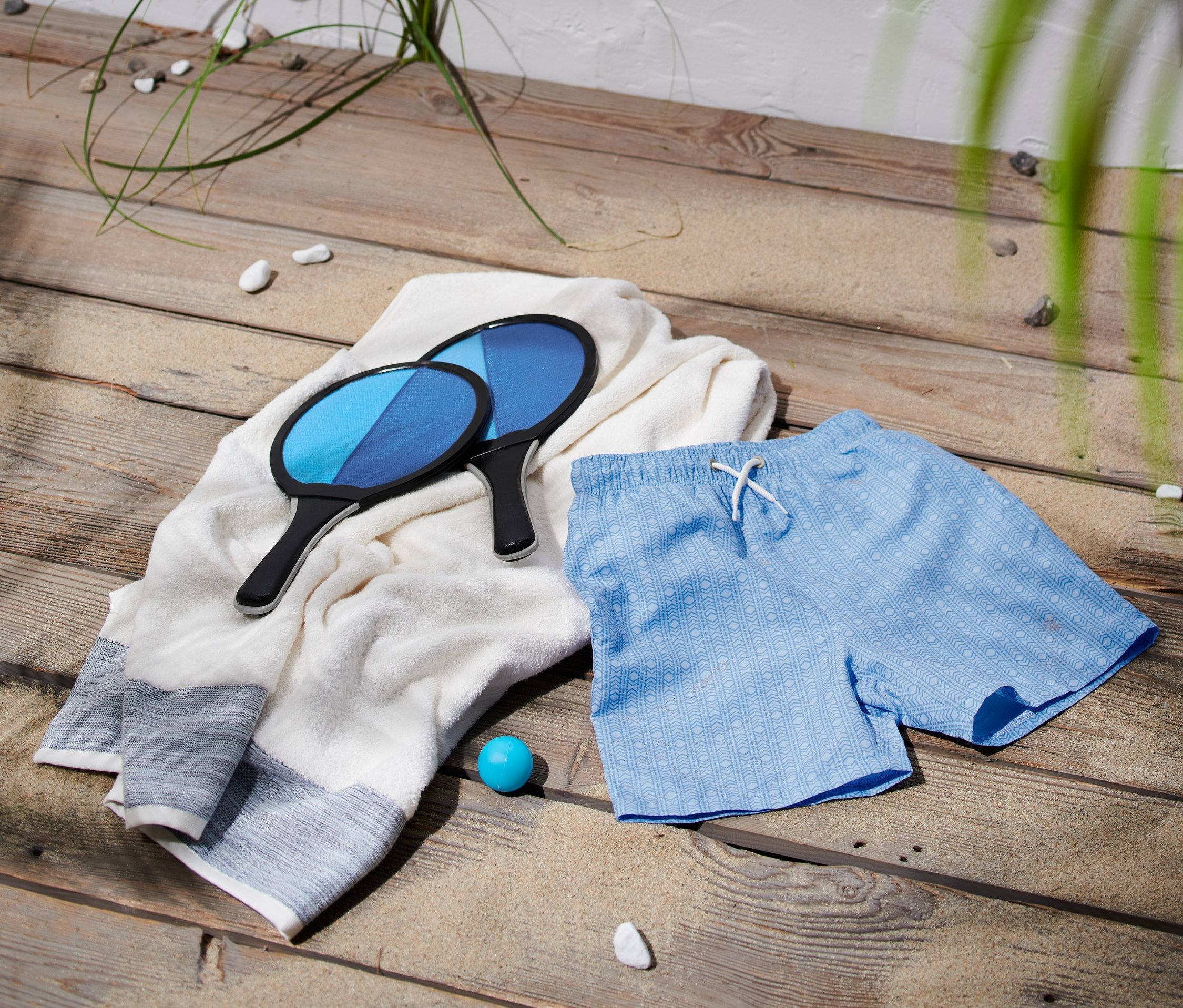 Deux raquettes de plage reposent sur une serviette blanche à côté d'un short de bain bleu en polyester recyclé.