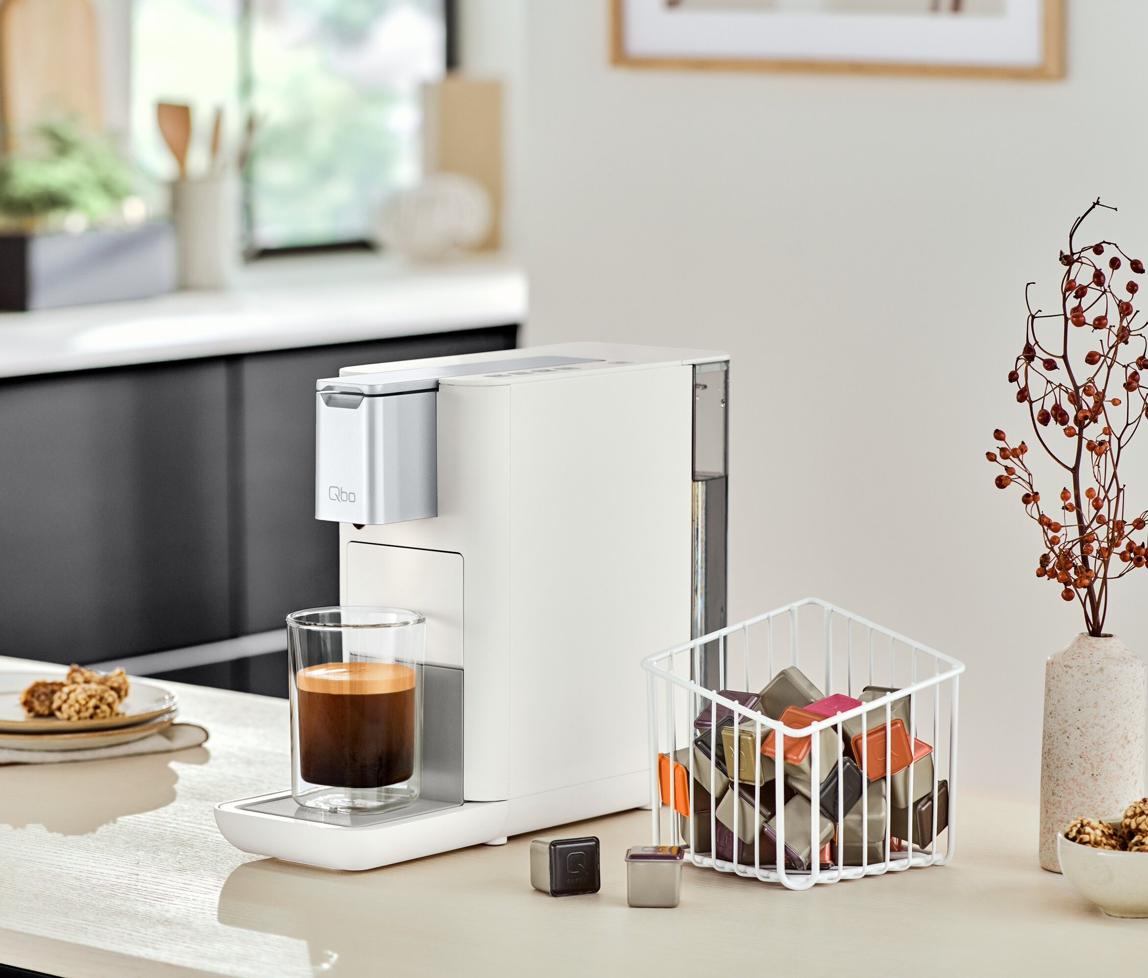 Une machine à café Qbo ESSENTIAL Bright Sand est placée à côté d'un verre de café sur un comptoir de cuisine.