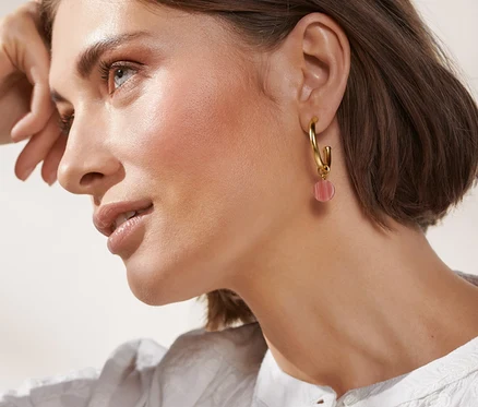 Une femme regarde vers le haut et porte des créoles dorées 23 carats avec une pierre synthétique et un chemisier blanc.