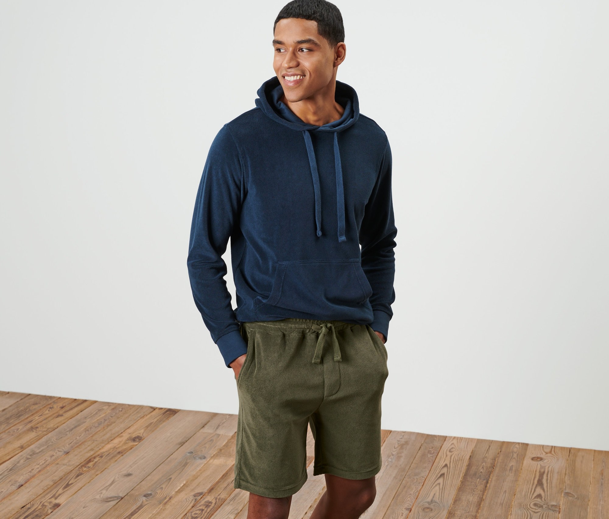 Un homme souriant se tient debout et porte un sweat à capuche bleu et un short en tissu éponge vert.