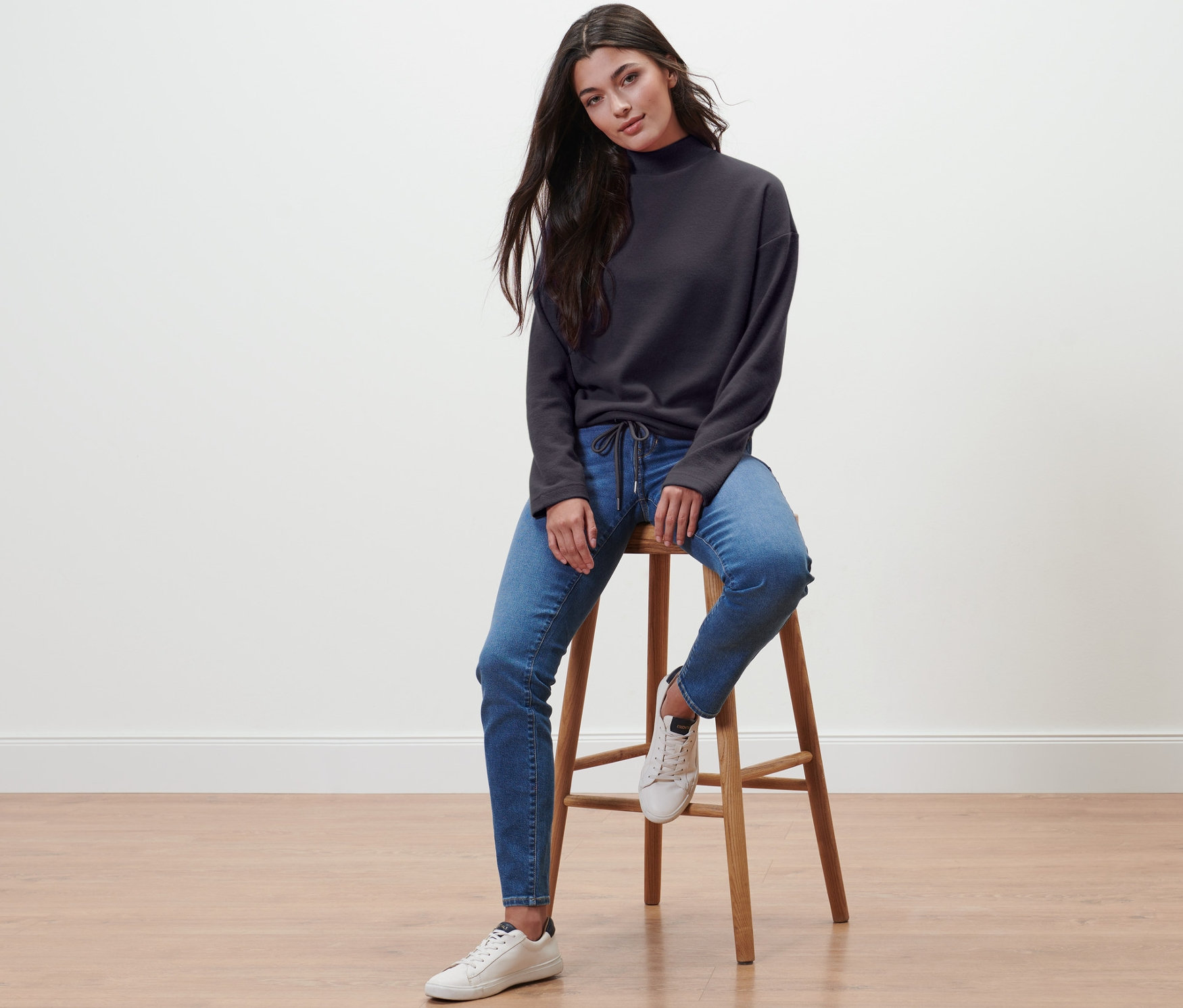 Une femme est assise sur un tabouret et porte un sweat-shirt avec col montant, un jean et des baskets.