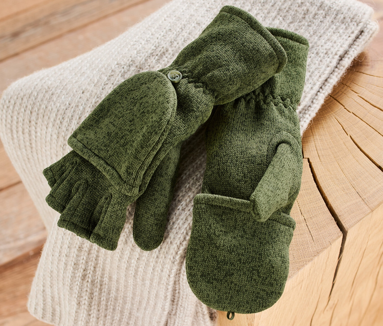 Des moufles convertibles en polaire tricotée kaki reposent sur un pull sur une table en bois.