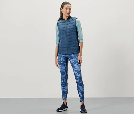 Une femme porte un gilet matelassé thermique, un legging bleu et des baskets foncées.