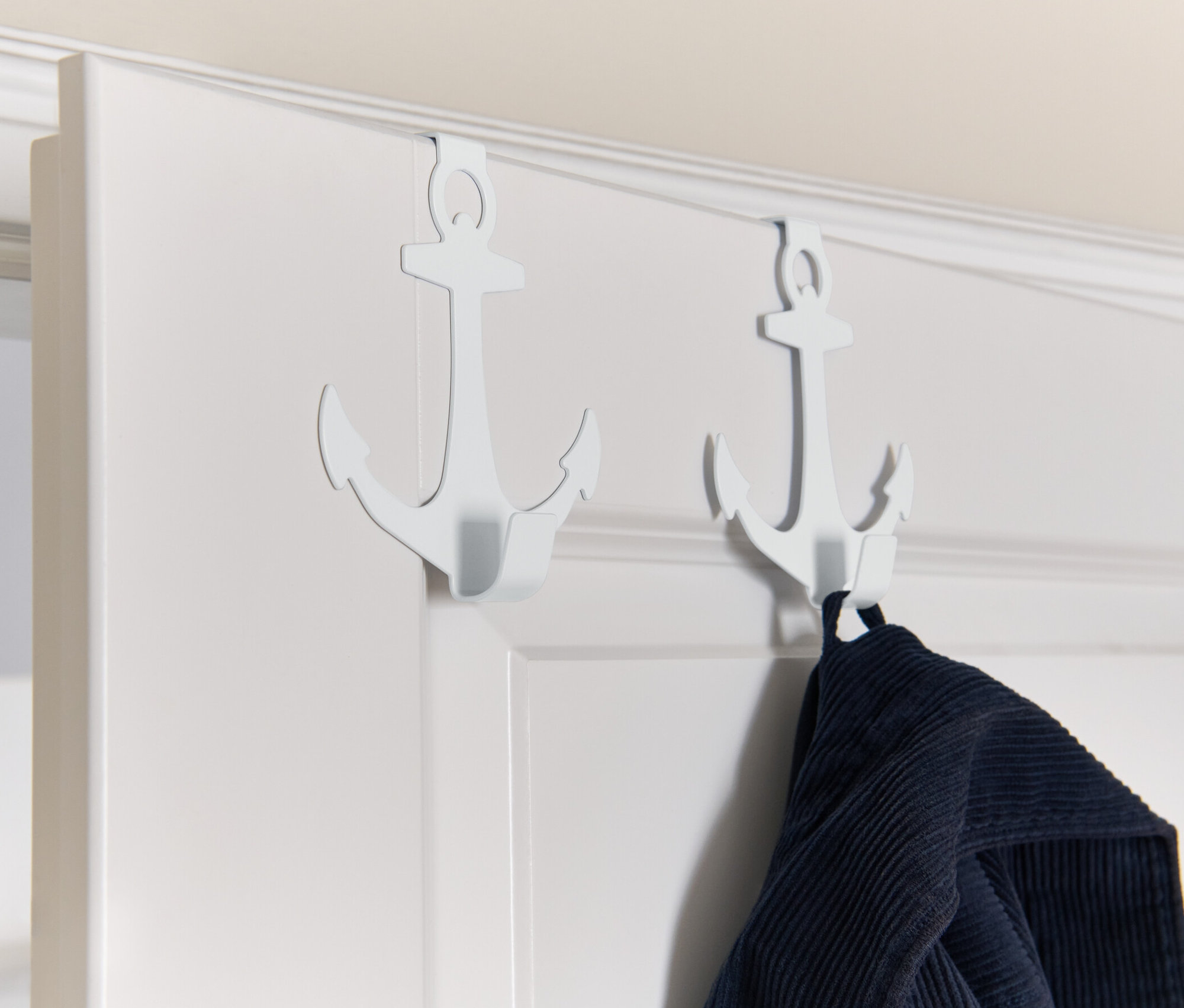 Deux crochets de porte blancs en forme d'ancre suspendus à une porte blanche, avec un vêtement bleu accroché à l'un d'eux.