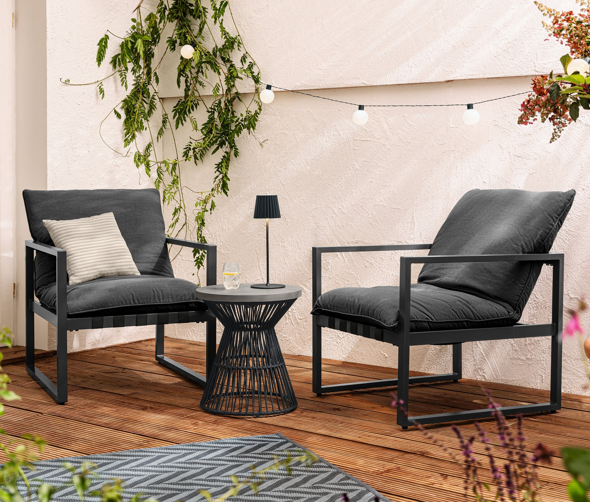 Deux fauteuils de relaxation sont placés sur une terrasse à côté d'une table d'appoint avec un plateau en polyciment. Une lampe de table noire à LED rechargeable est posée sur la table. Une guirlande lumineuse d'extérieur à LED est suspendue au-dessus des fauteuils.