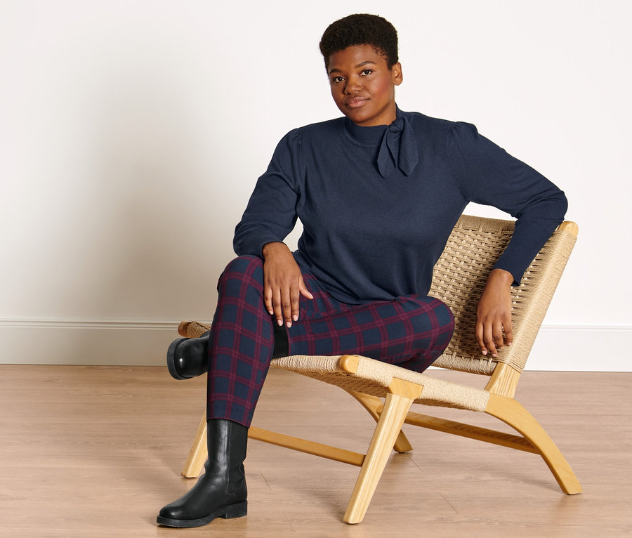 Une femme est assise sur une chaise. Elle porte un pull en maille fine avec un nœud, un pantalon stretch à carreaux rouges et bleus et des bottines en cuir.