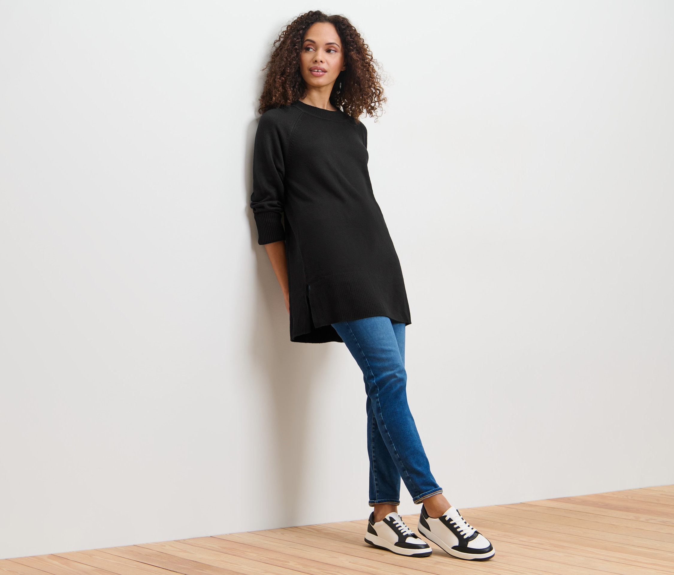 Une femme appuyée contre un mur portant un long pull en maille noir.