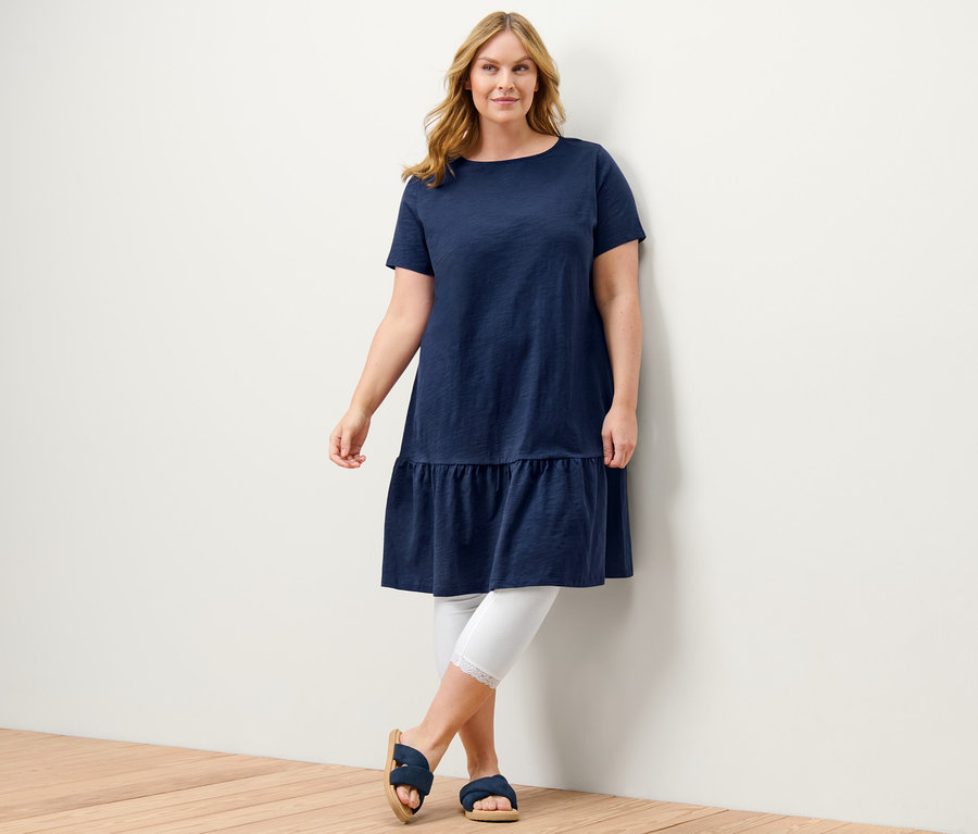 Une femme pose en robe en jersey à volants bleu foncé et legging 3/4 blanc avec bordure en dentelle.