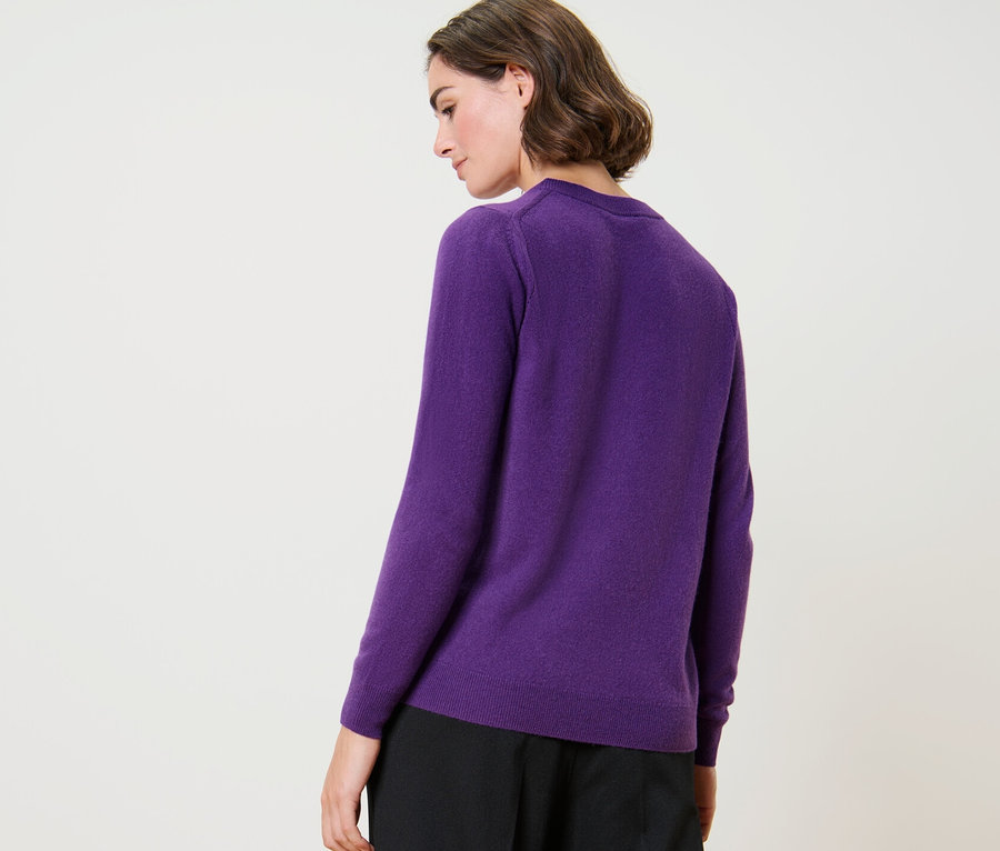 Une femme pose dos à la caméra et porte un pull violet en maille fine avec du cachemire et un pantalon noir.