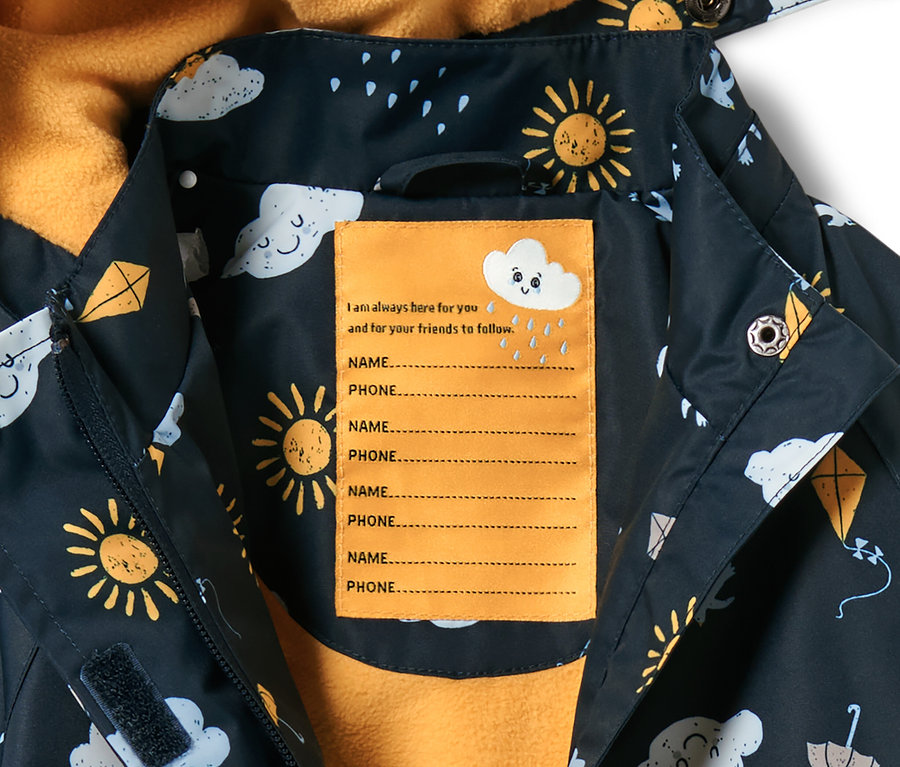 Gros plan sur une veste de pluie thermique bleu foncé pour tout petit, avec imprimé intégral de soleil, nuages et cerfs-volants. Une étiquette jaune pour le nom et le numéro de téléphone se trouve à l'intérieur.
