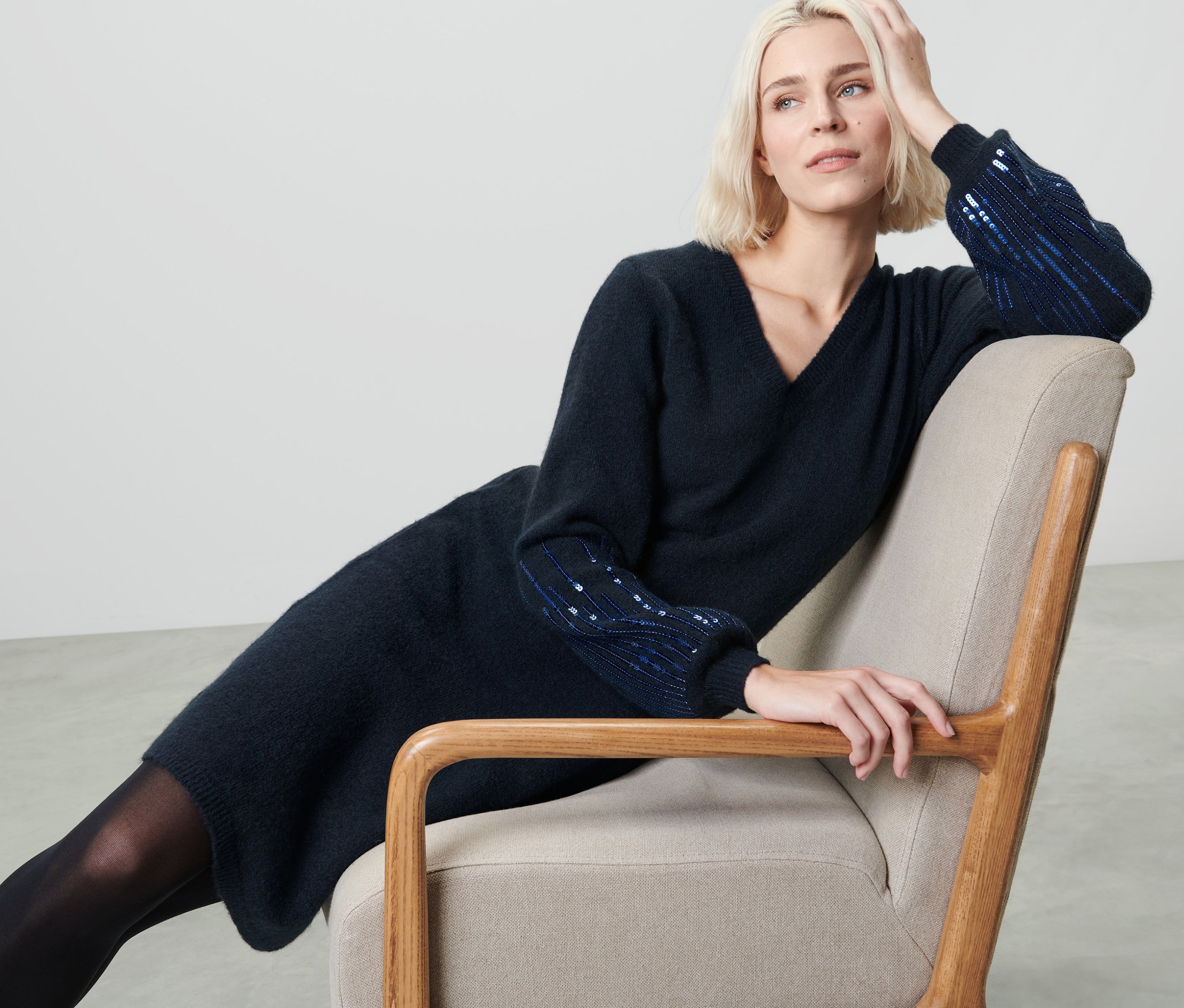 Une femme blonde est assise dans un fauteuil et porte une robe en maille bleu foncé à paillettes.