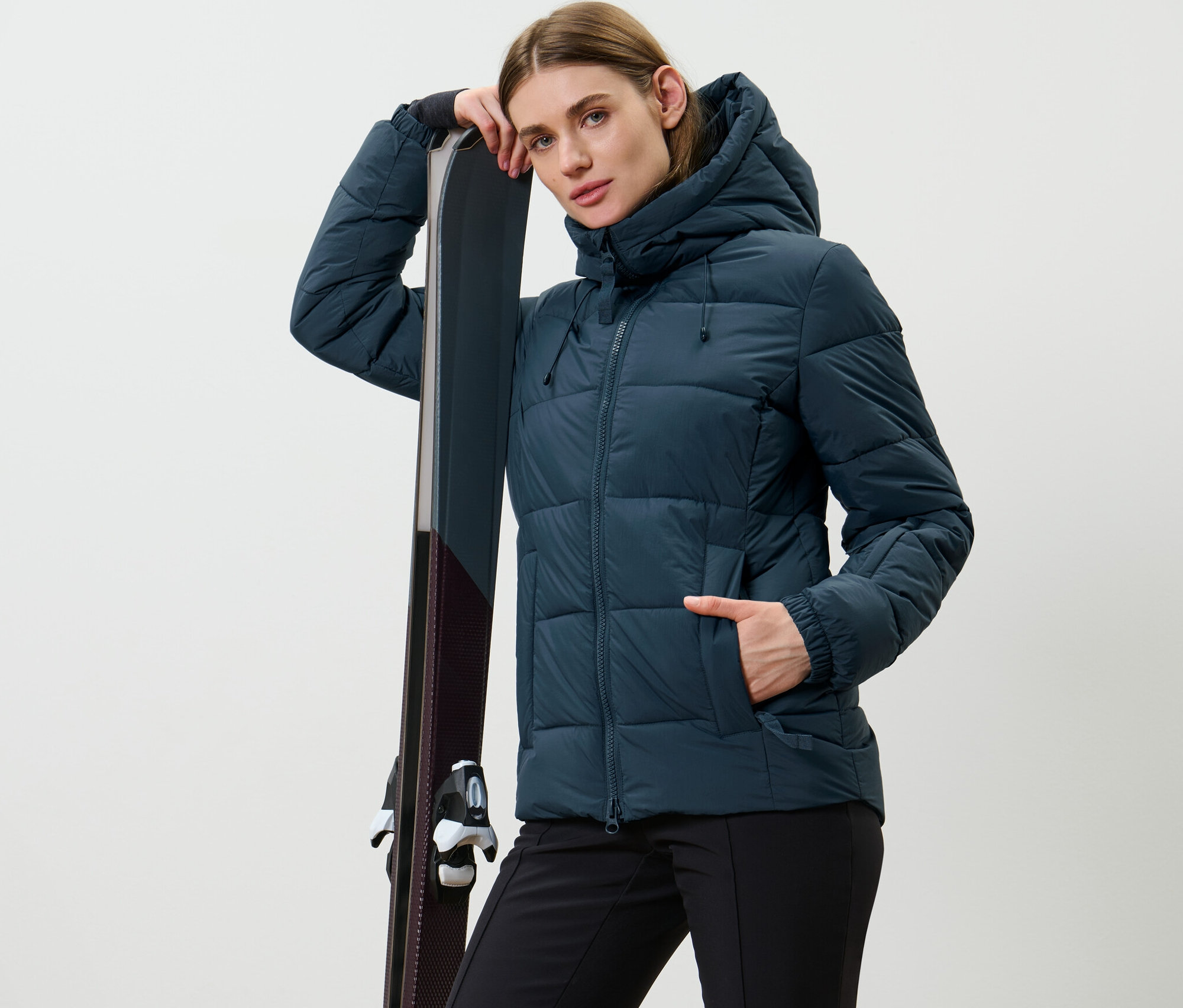 Femme posant en veste de ski matelassée bleu marine, appuyée sur des skis.