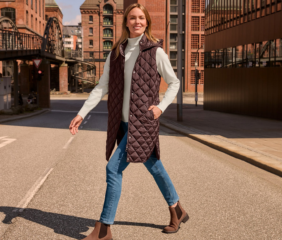 Une femme marche dans la rue et porte un long gilet sans manches matelassé avec capuche, un pull à col roulé en maille fine, blanc crème, un jean slim – Coupe «
Emma » bleu moyen et des bottines en cuir velours.
