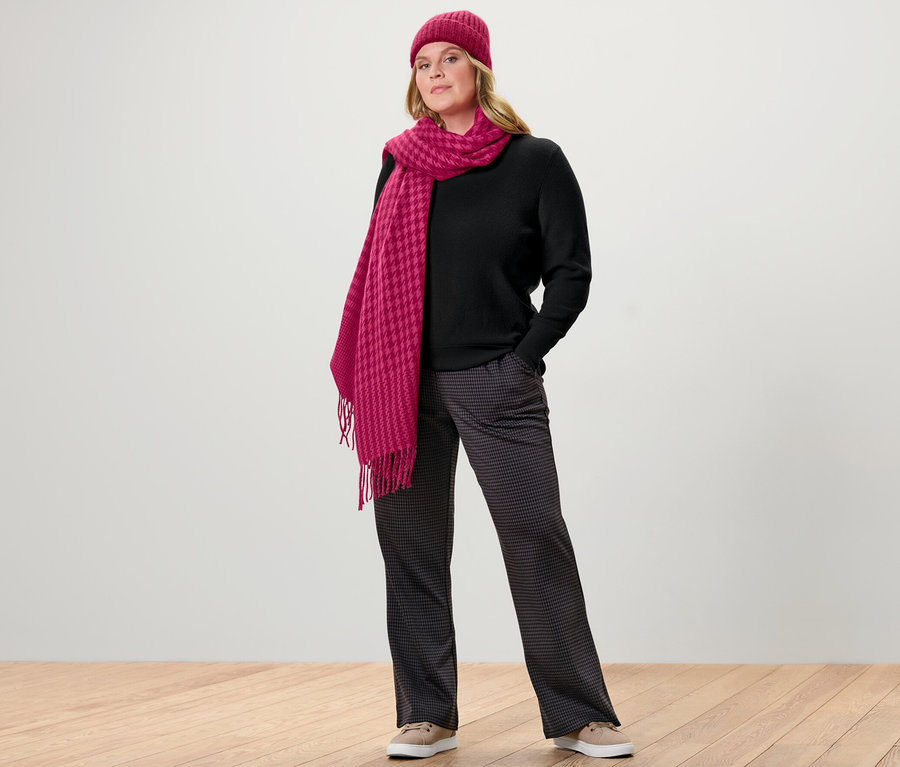 Femme posant en pull en maille fine, noir, et pantalon à enfiler à jacquard pied-de-poule. Elle porte un bonnet et une écharpe roses.