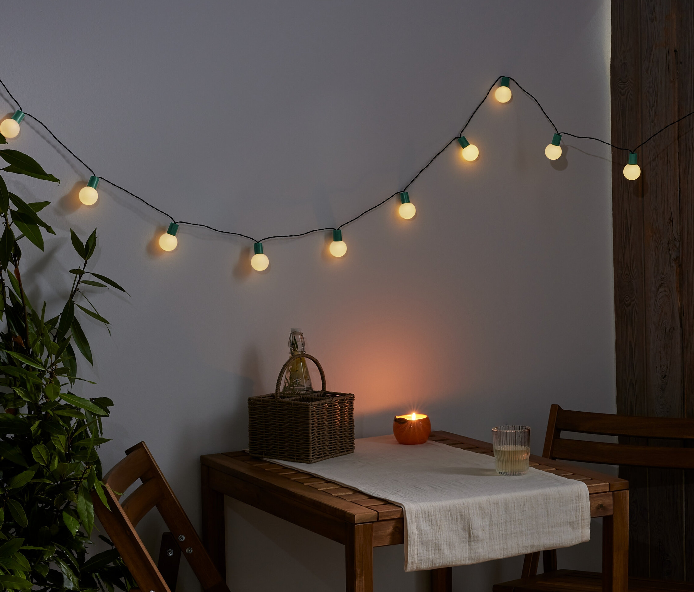 Une guirlande lumineuse à LED est accrochée au mur. Sur une table, il y a un panier, une bougie et un verre.