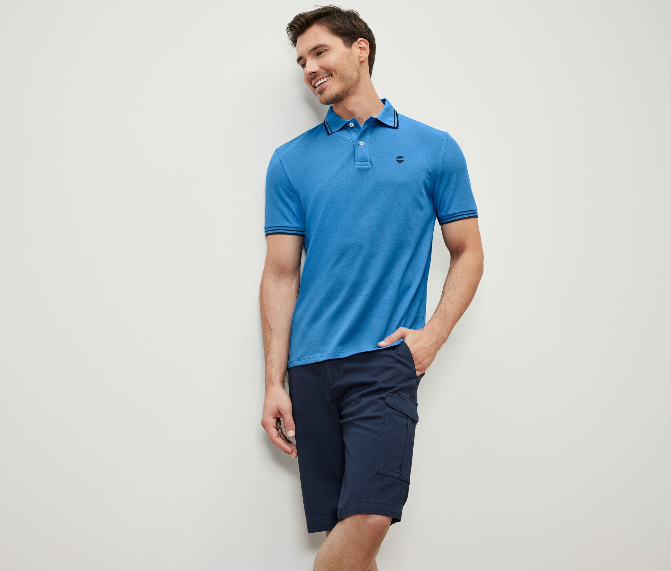 Un homme appuyé contre un mur porte un polo en piqué bleu et un short bleu marine.