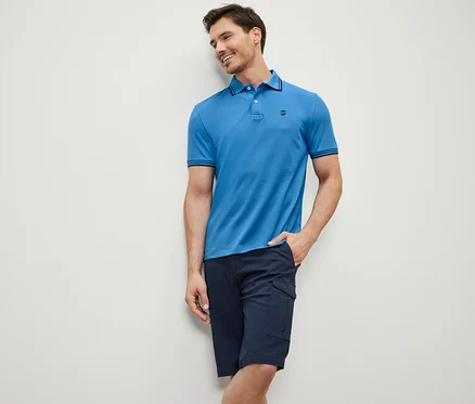 Un homme appuyé contre un mur porte un polo en piqué bleu et un short bleu marine.