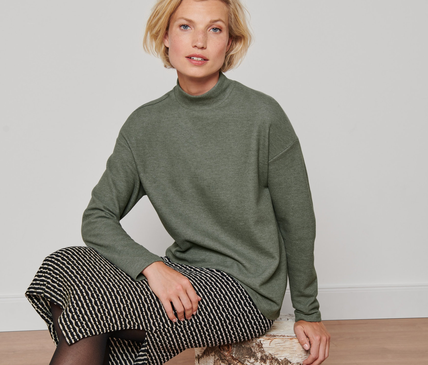 Une femme est assise sur une souche d'arbre et porte un t-shirt manches longues avec col montant vert, une jupe noire et des bottes noires.