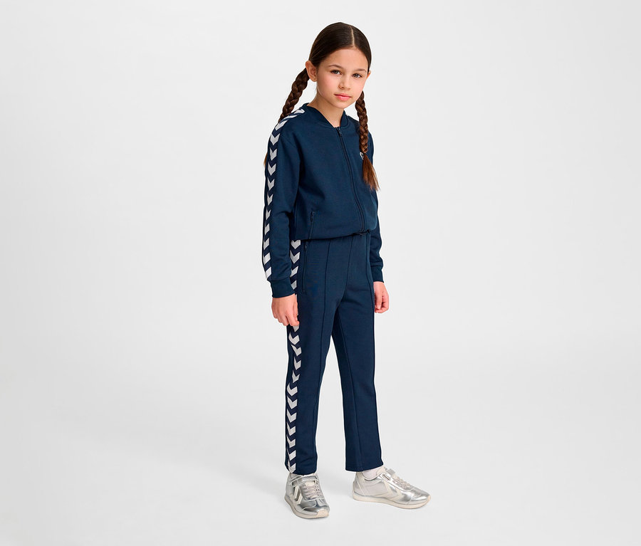 Une fille avec des tresses portant un survêtement bleu marine Hummel HMLJR Archive Regular Poly Pants et des chaussures argentées se tient sur un fond clair.