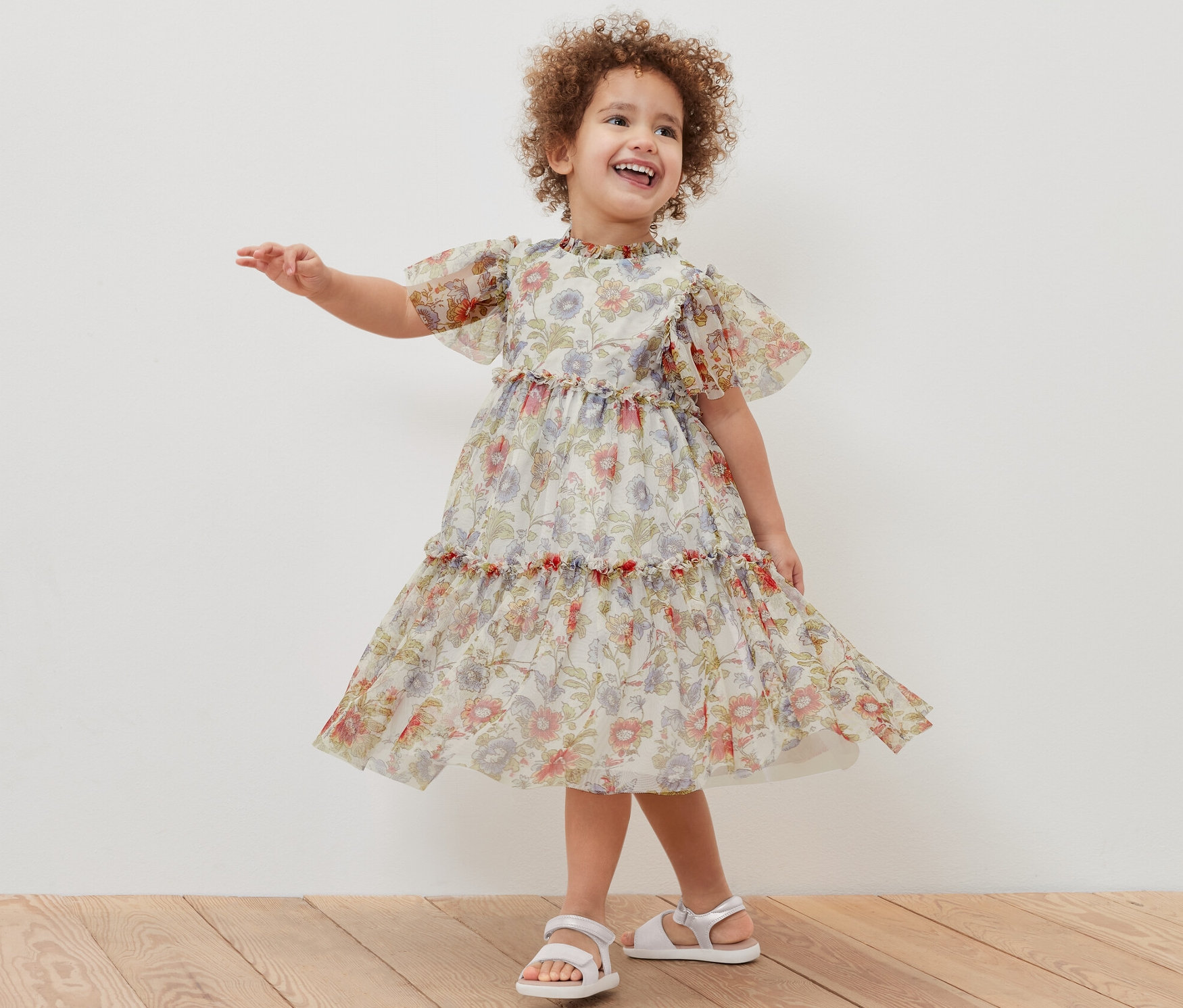 Une fillette souriante pose dans une robe en tulle à fleurs et des sandales blanches.
