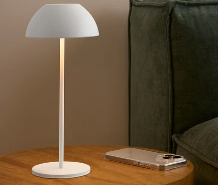 Une lampe blanche est posée sur une table en bois à côté d'un téléphone portable et d'un canapé vert.