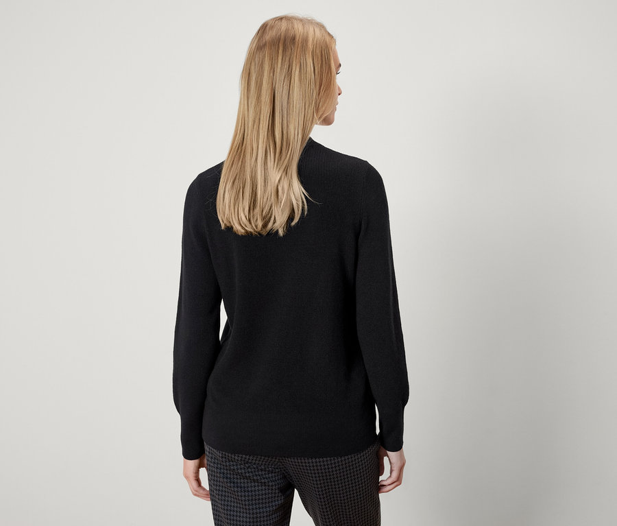 Vue de dos, une femme aux cheveux blonds porte un pull noir et un pantalon à motifs.