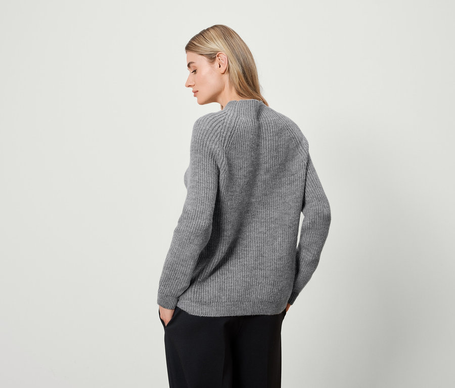 Une femme blonde portant un pull gris et un pantalon noir se tient dos à l'objectif.