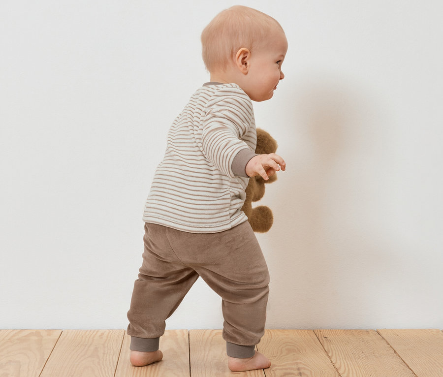 Un bébé dans un ensemble en nicki tient un ours en peluche sur un plancher en bois.