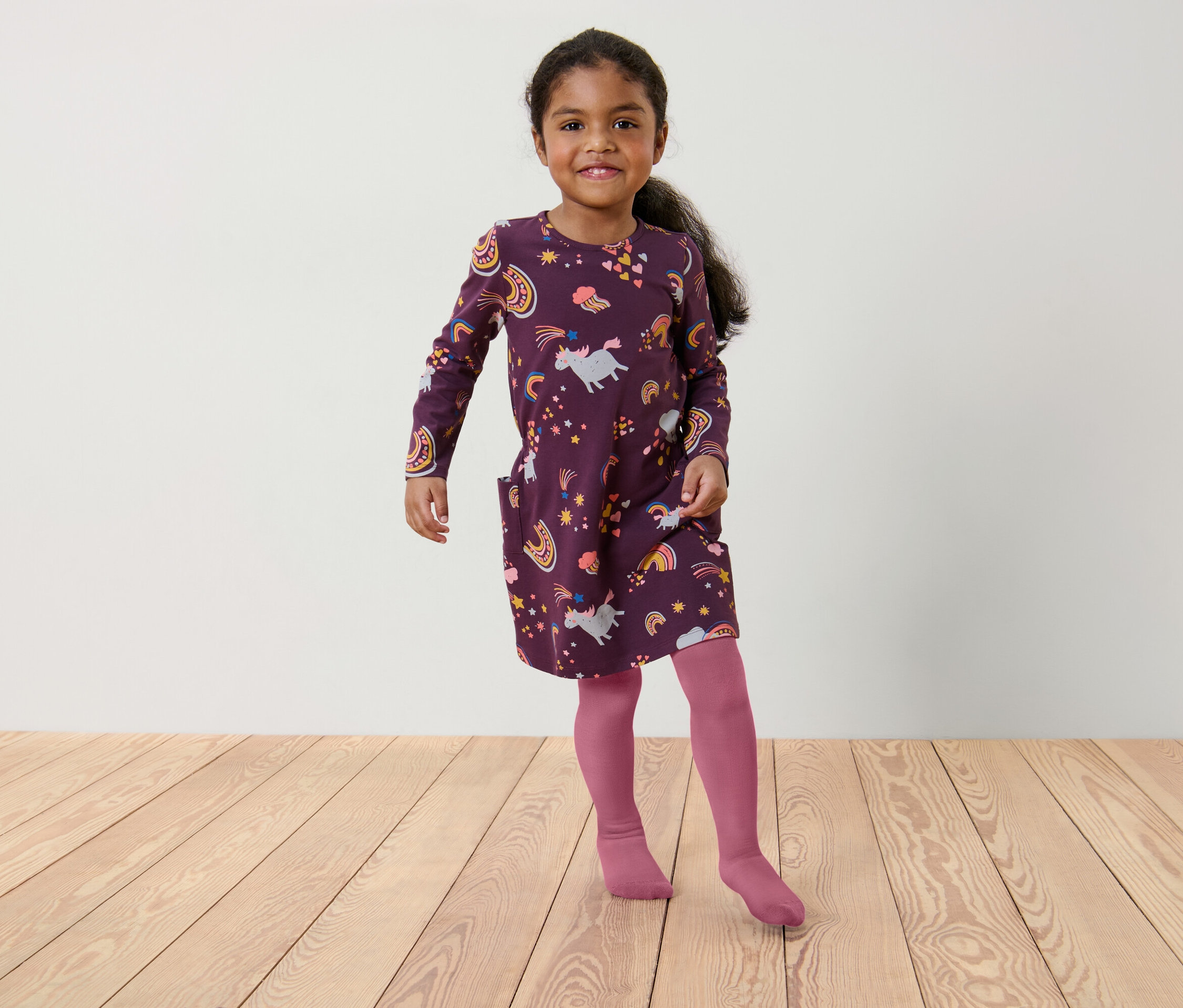 Une fillette portant une robe bordeaux à motifs de licornes et d'arcs-en-ciel et des collants roses se tient sur un plancher en bois.