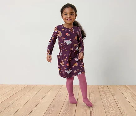 Une fillette portant une robe bordeaux à motifs de licornes et d'arcs-en-ciel et des collants roses se tient sur un plancher en bois.