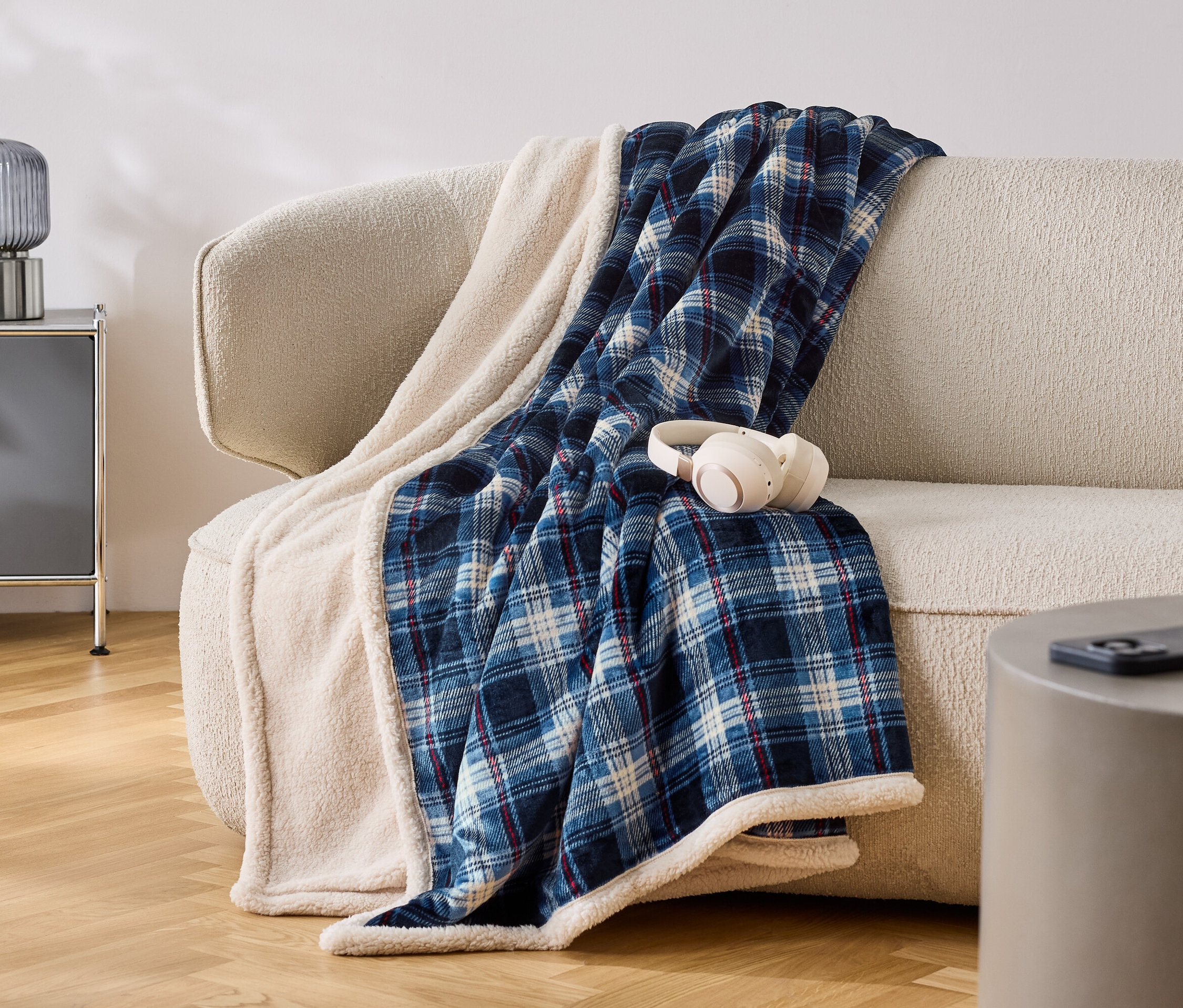 Sur un canapé beige est posée une couverture à carreaux bleu et blanc avec une bordure blanche. Des écouteurs blancs sont posés sur la couverture.