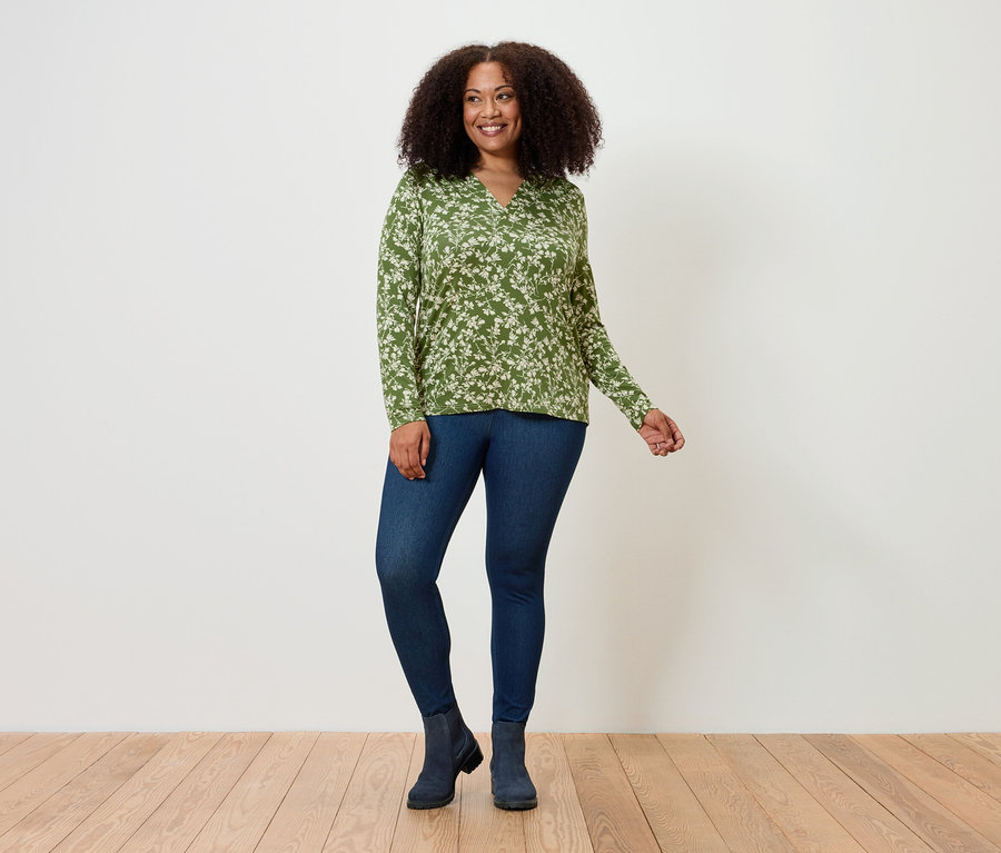 Une femme se tient debout et pose sur un plancher en bois, elle porte un haut vert à motifs floraux, un legging en jean et des bottes.