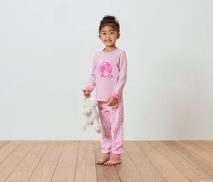 Une fille en pyjama rose Barbie tenant un ours en peluche.