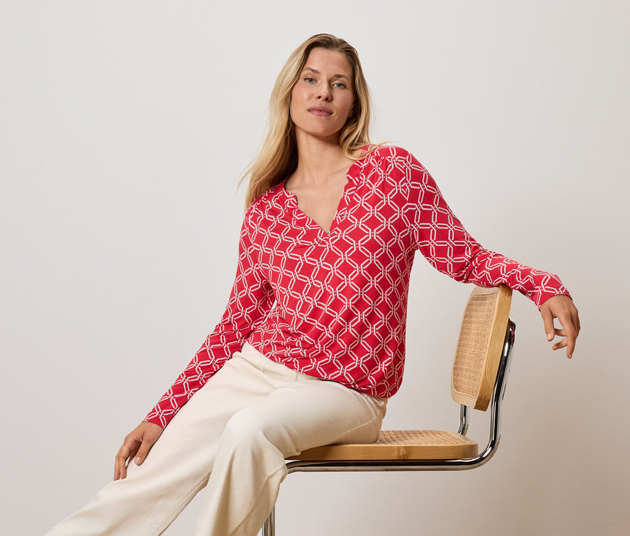 Femme blonde assise sur une chaise, portant un haut rouge et blanc et un pantalon crème.