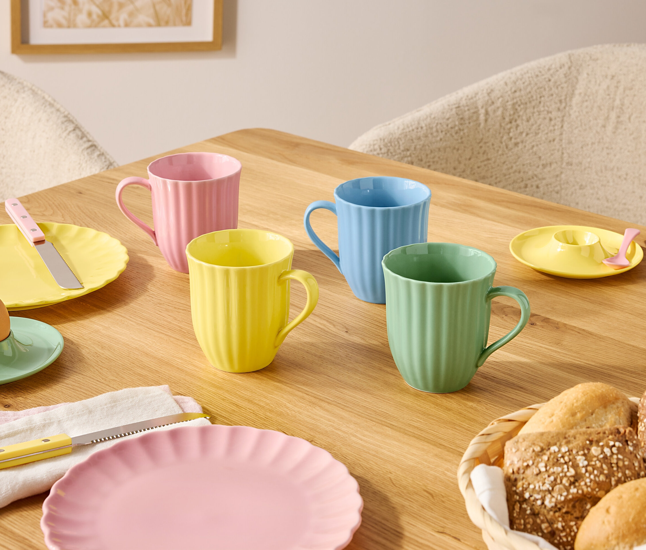 Sur une table en bois sont disposées des assiettes et des tasses de couleurs pastel ainsi qu'une corbeille à pain.