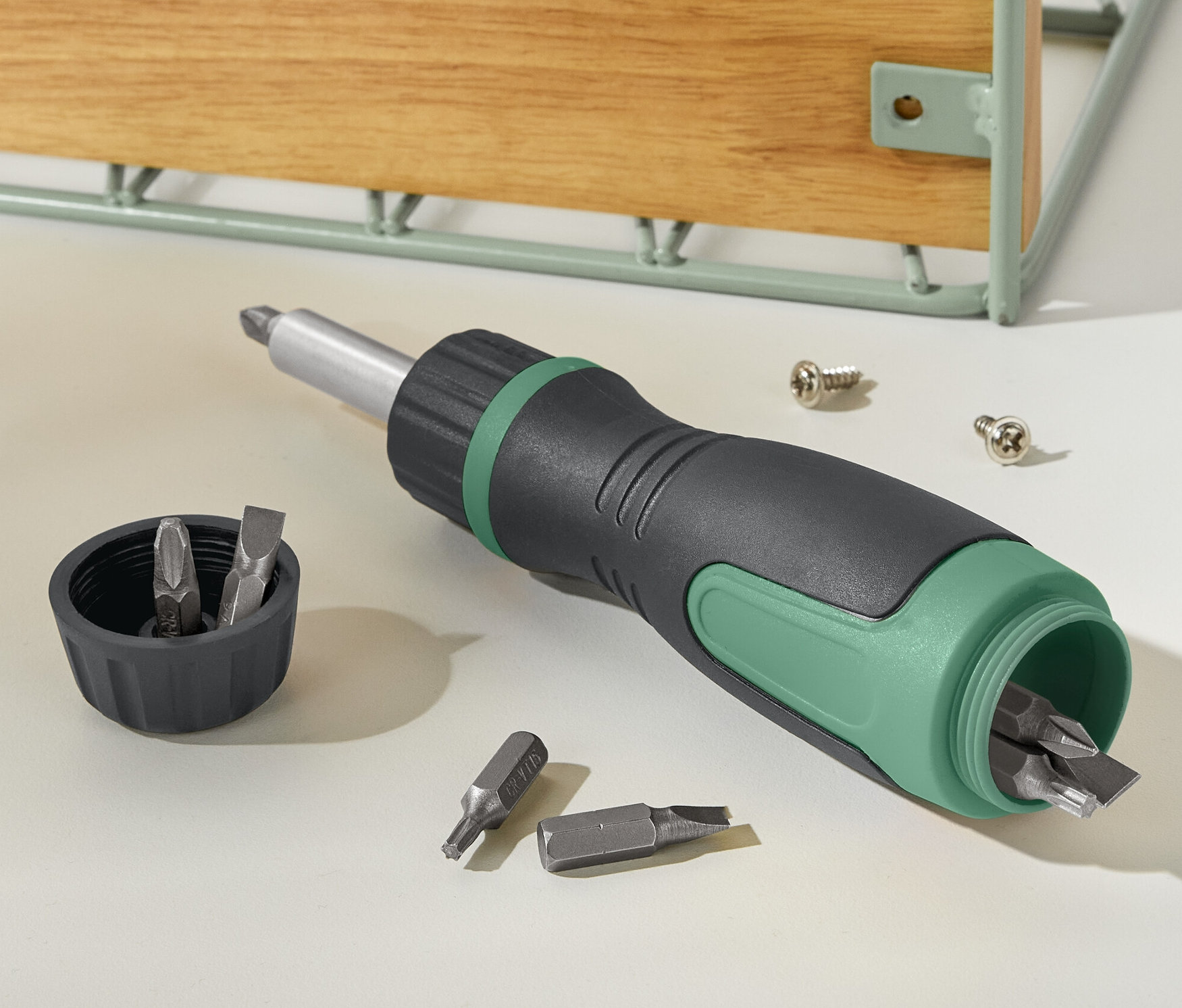 Un tournevis à cliquet magnétique noir et vert repose à côté d'un couvercle avec des embouts et de deux vis.