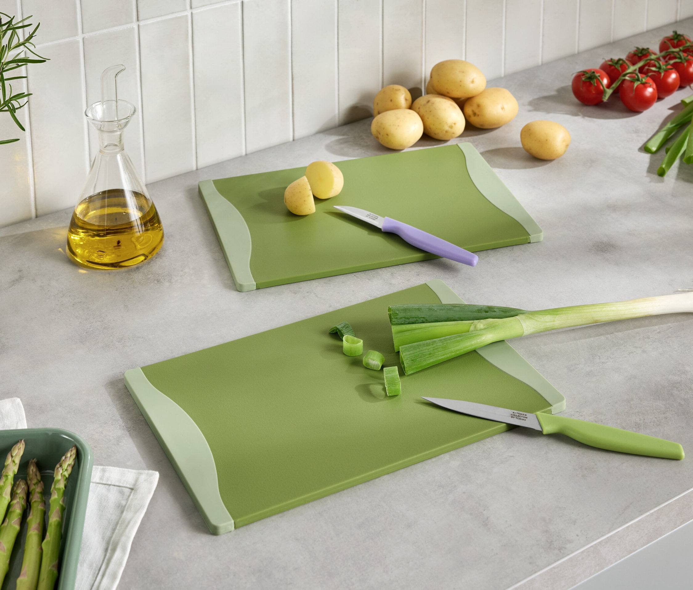Deux planches à découper vertes avec des légumes coupés, des couteaux, des pommes de terre, des tomates, des poireaux, des asperges et de l'huile sur un comptoir de cuisine.