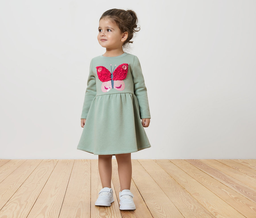 Une fillette en robe verte avec un papillon se tient sur un plancher en bois.