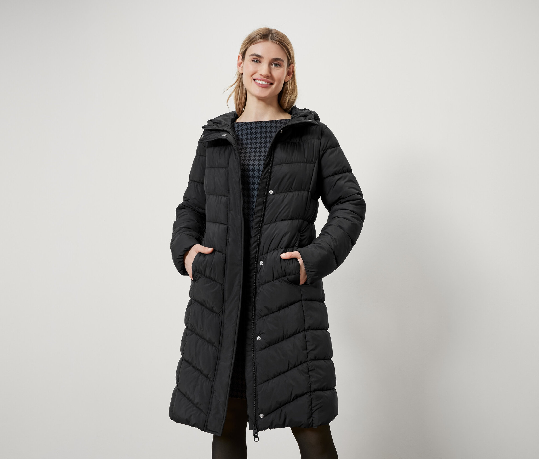 Une femme aux cheveux blonds se tient debout et porte un long manteau noir matelassé avec une capuche.