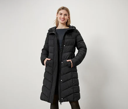 Une femme aux cheveux blonds se tient debout et porte un long manteau noir matelassé avec une capuche.