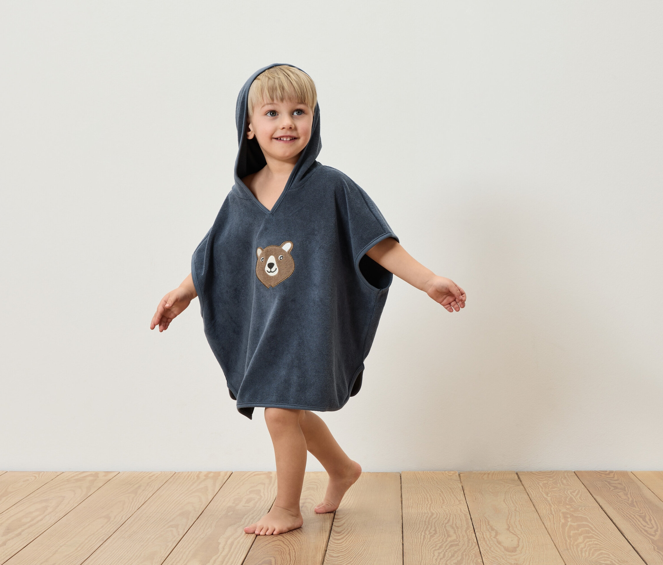 Un garçon portant un poncho bleu à capuche avec un motif d'ours debout sur un plancher en bois devant un mur blanc.