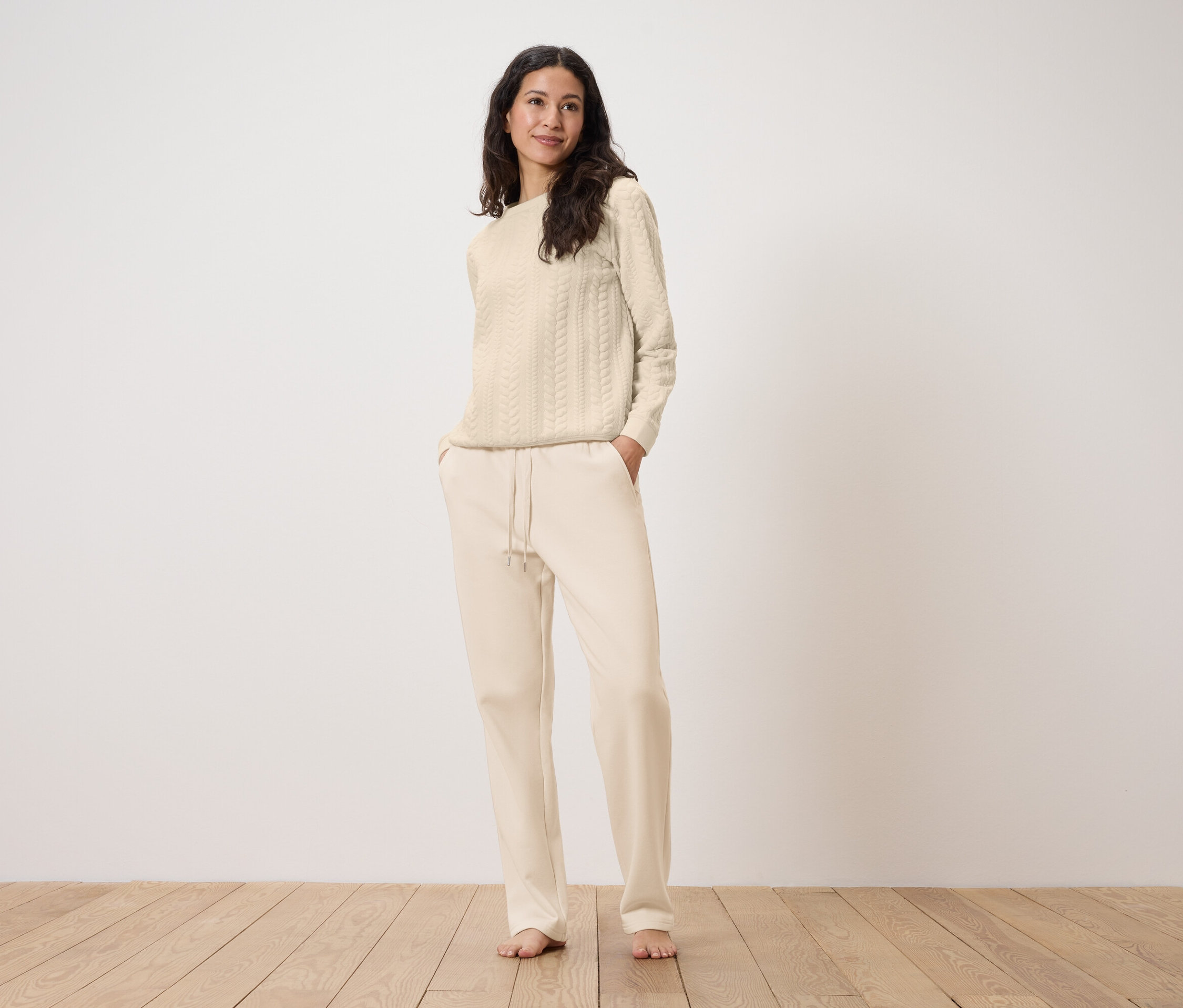 Une femme se tient debout, les mains dans les poches d'un pantalon de survêtement crème avec un pull crème à torsades.