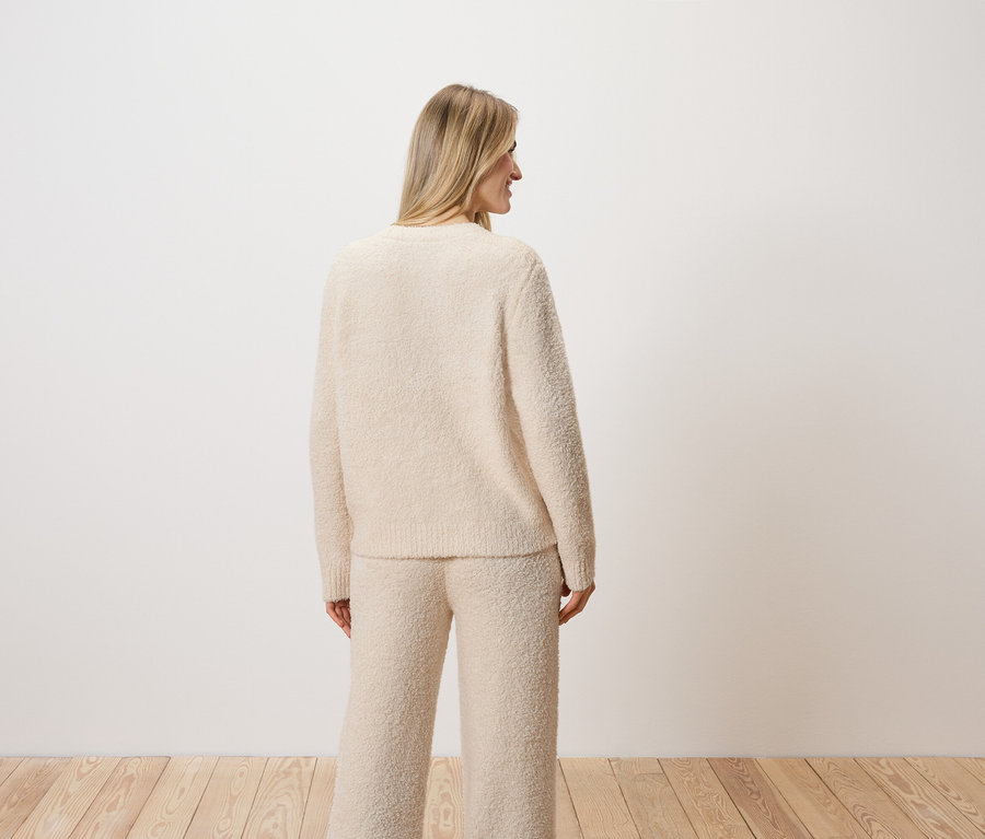 Une femme blonde se tient dos à la caméra et porte un pull et un pantalon couleur crème.