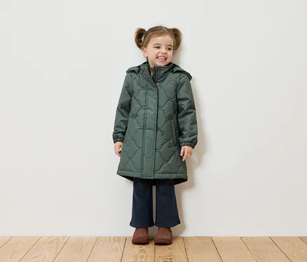 Une fillette souriante avec des couettes se tient devant un mur blanc et porte un manteau vert, un pantalon bleu marine et des chaussures marron.