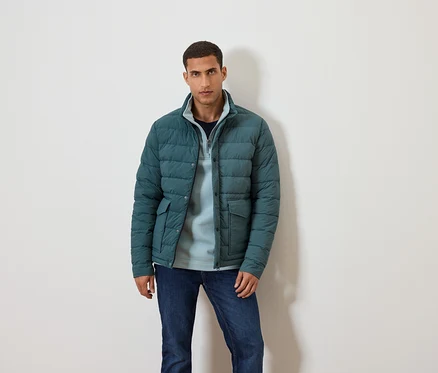 Un homme pose sur un fond blanc vêtu d'une veste bleue, d'un pull bleu et d'un jean.