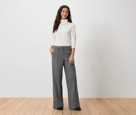 Une femme se tient debout, les mains dans les poches d'un pantalon gris, portant un col roulé blanc.