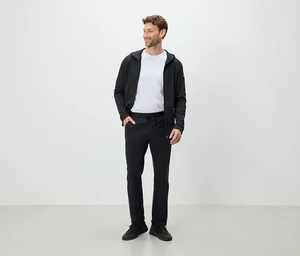 Un homme debout regardant sur le côté, portant un pantalon noir, un t-shirt blanc et un sweat à capuche noir.